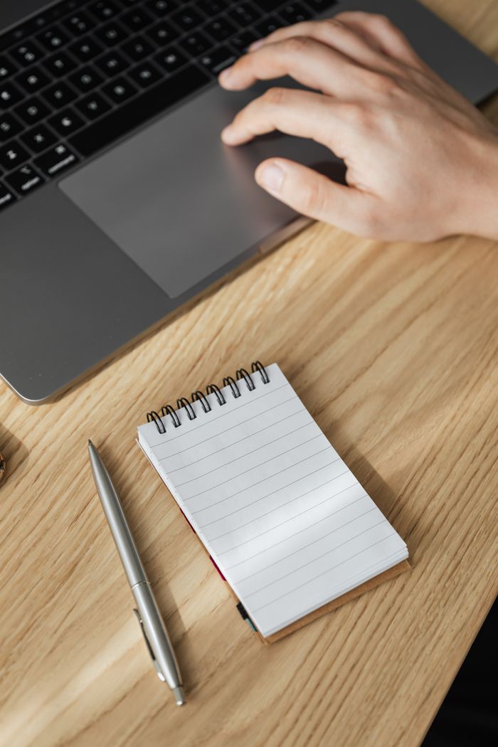 From above of crop anonymous male working on netbook touchpad placed on wooden table with ring bound notebook and stylish silver pen
